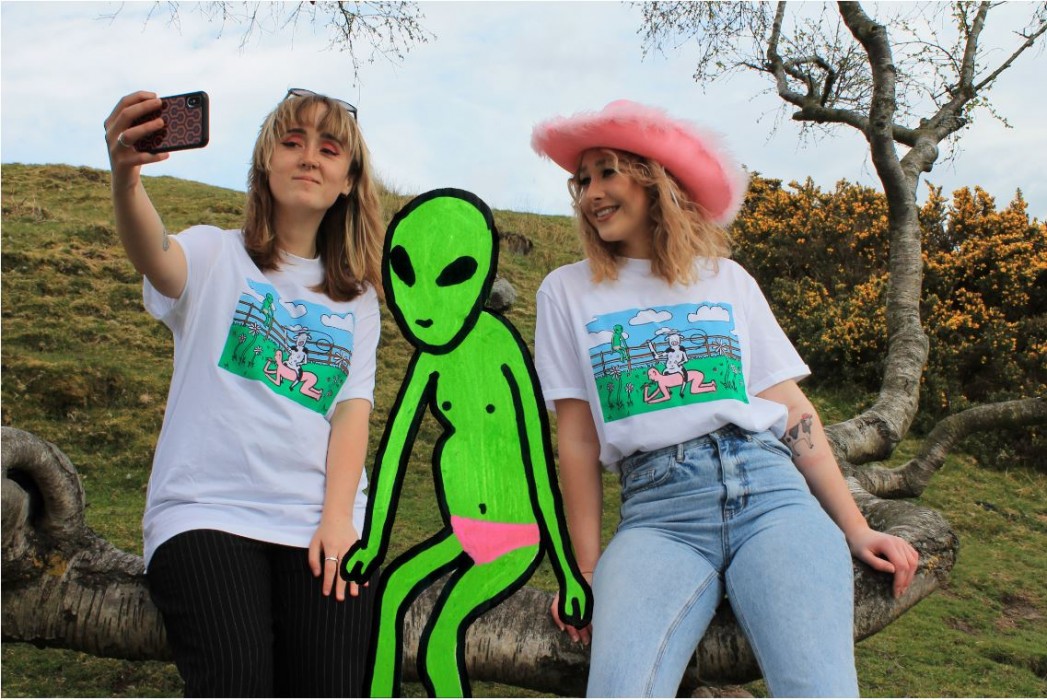 A advert showing two women modelling T-shirt designs, taking a seflie with a green cartoon alien.