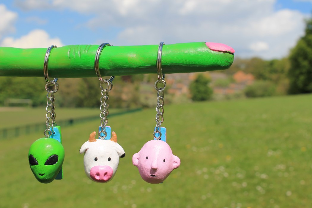 Three keyrings in the shape of heads. Characters include an alien, a cow, and a man. Displayed on a green alien finger